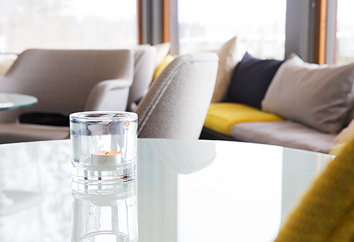 Kahvilan lasipöytä, jolla tuikkukynttilä lasisessa tuikkukupissa. Vaaleat tuolit, taustalla ikkunapenkkien keltaisia, tummansinisiä ja vaaleita tyynyjä. Kuva on valoisa ja välittää kahvilan modernin tunnelman.